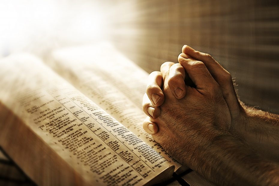 Hands clasped in prayer over an open Bible with light shining from above.