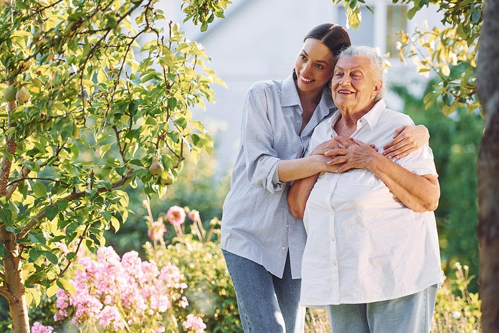 A younger woman embraces an older woman from behind as they both smile, standing in a lush garden with blooming flowers and green trees, in the serene environment of an assisted living community.