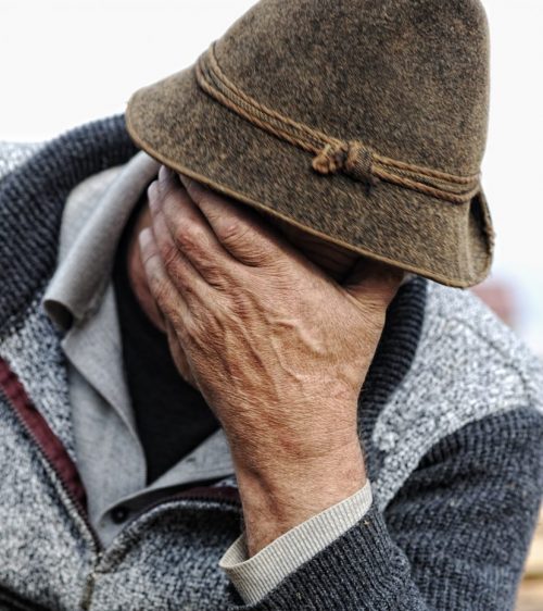 Person wearing a brown hat and grey jacket, covering their face with their right hand.