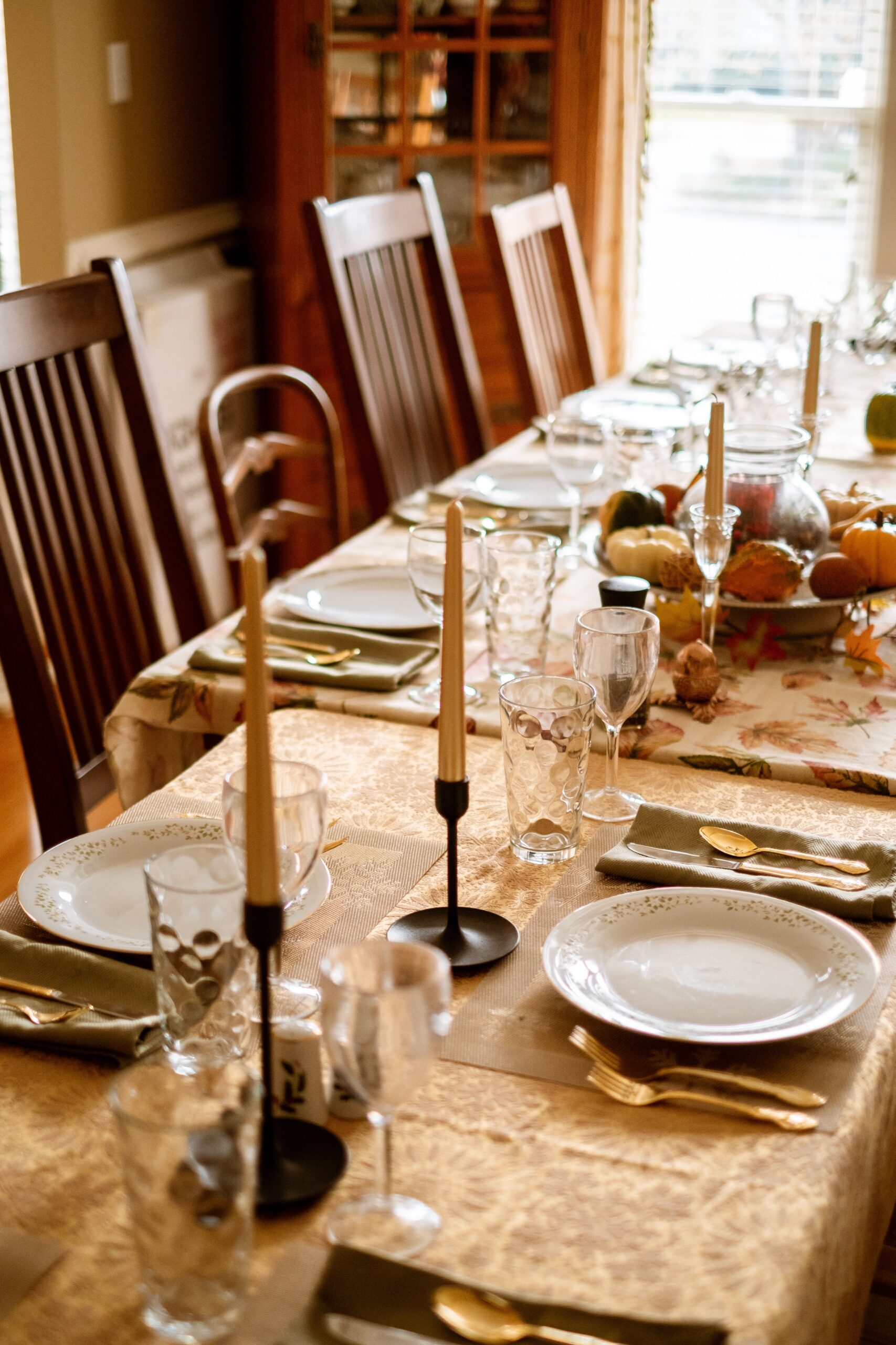 A dining table is set for a meal with chairs around it. The table has plates, glasses, cutlery, candles, and a centerpiece with fruits and vegetables.