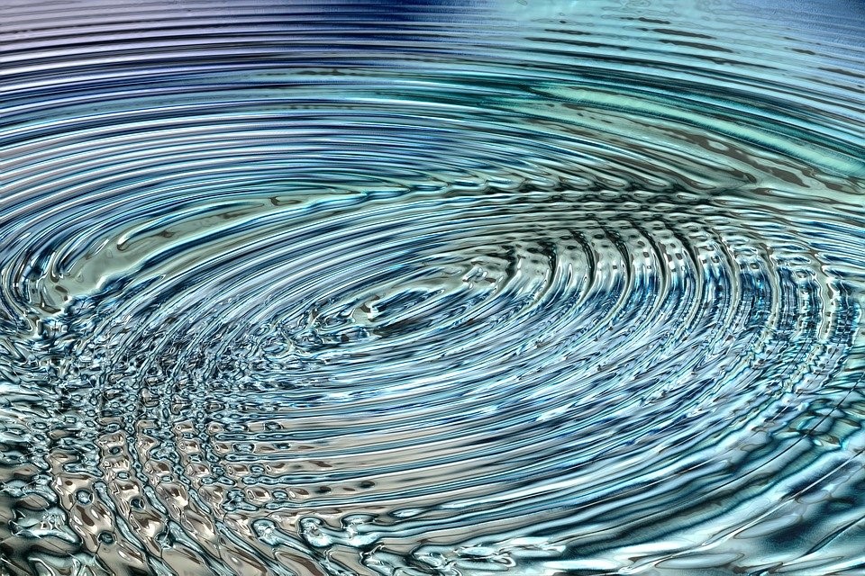 Close-up of water surface with concentric ripples, creating a circular pattern. The water has a clear, reflective quality with blue and green hues.