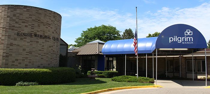 Entrance of Pilgrim Manor featuring a blue-canopied walkway and an American flag, adjacent to a round brick structure labeled Hanish Memorial Chapel.