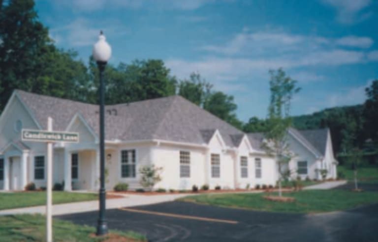 A single-story building with white walls and a gray roof sits on a landscaped area in Moundsville, West Virginia. A street sign reads "Candlewick Lane," and a lamppost is visible in the foreground. Trees are in the background, adding charm to this affordable housing community.