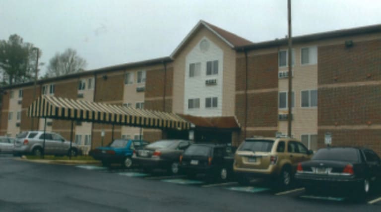 A three-story brick building with a covered entrance, designated as affordable housing in Winder, Georgia. Several cars are parked in a lot in front of the building under an overcast sky.