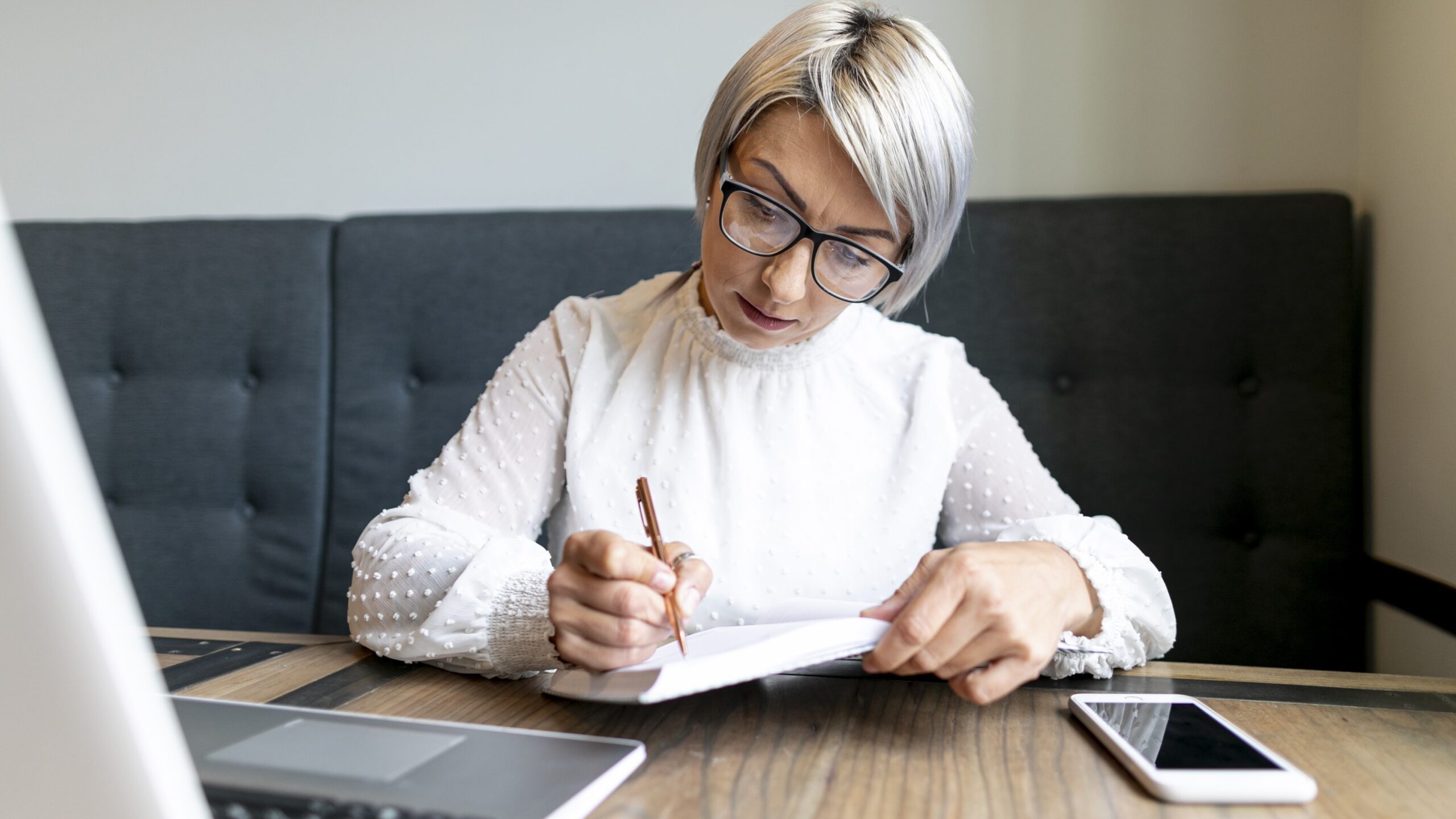 A woman with short grey hair and glasses is sitting at a table, writing on a piece of paper. A laptop and a smartphone are placed on the table.