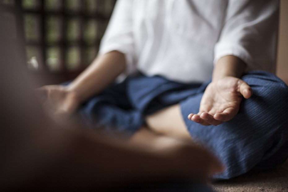 Person seated cross-legged on the floor, wearing a white shirt and blue pants, with hands resting on their knees, palms up, in a meditative posture.